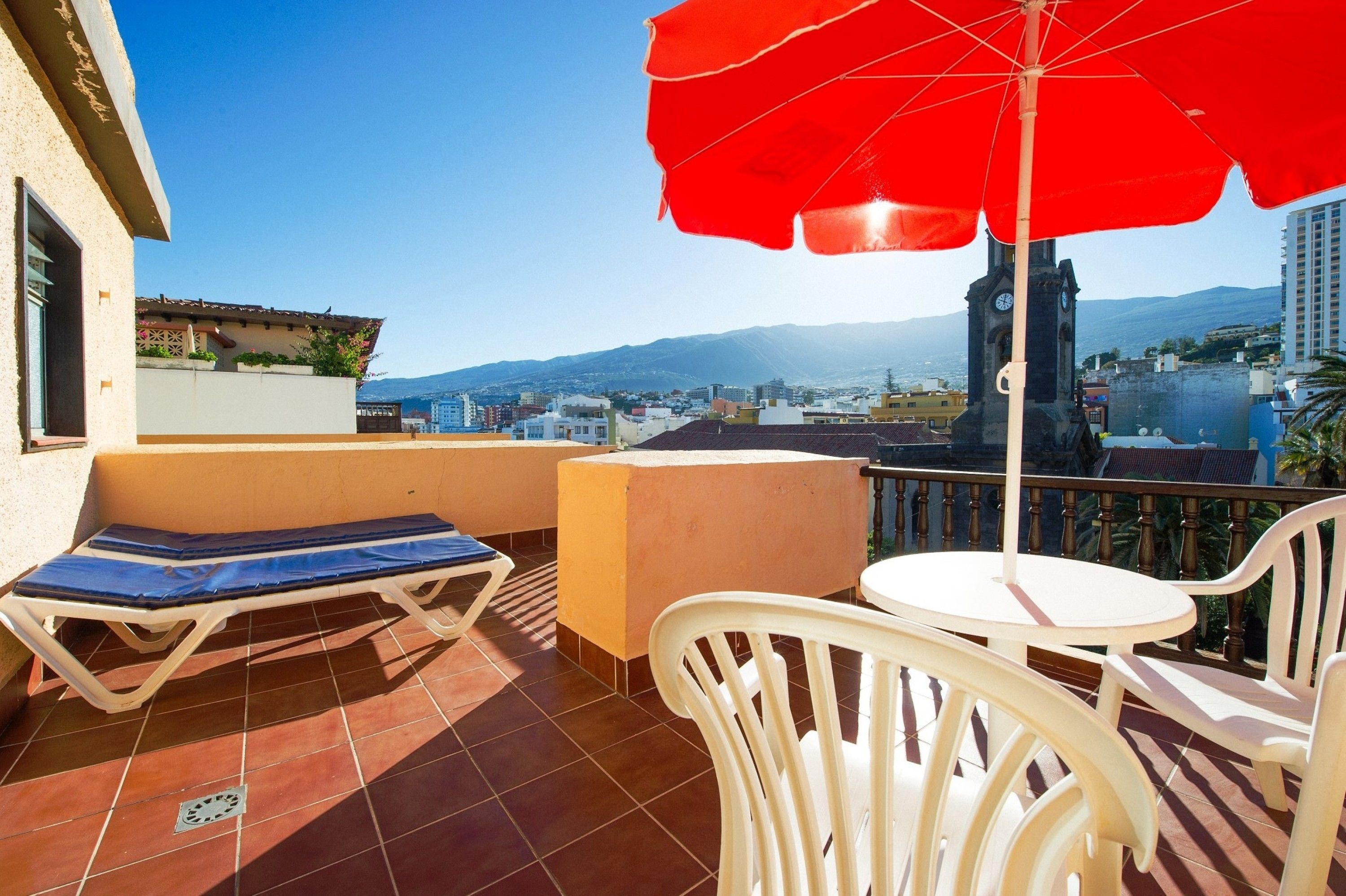 Ein Balkon mit rotem Sonnenschirm, einer Liege und einem Tisch.