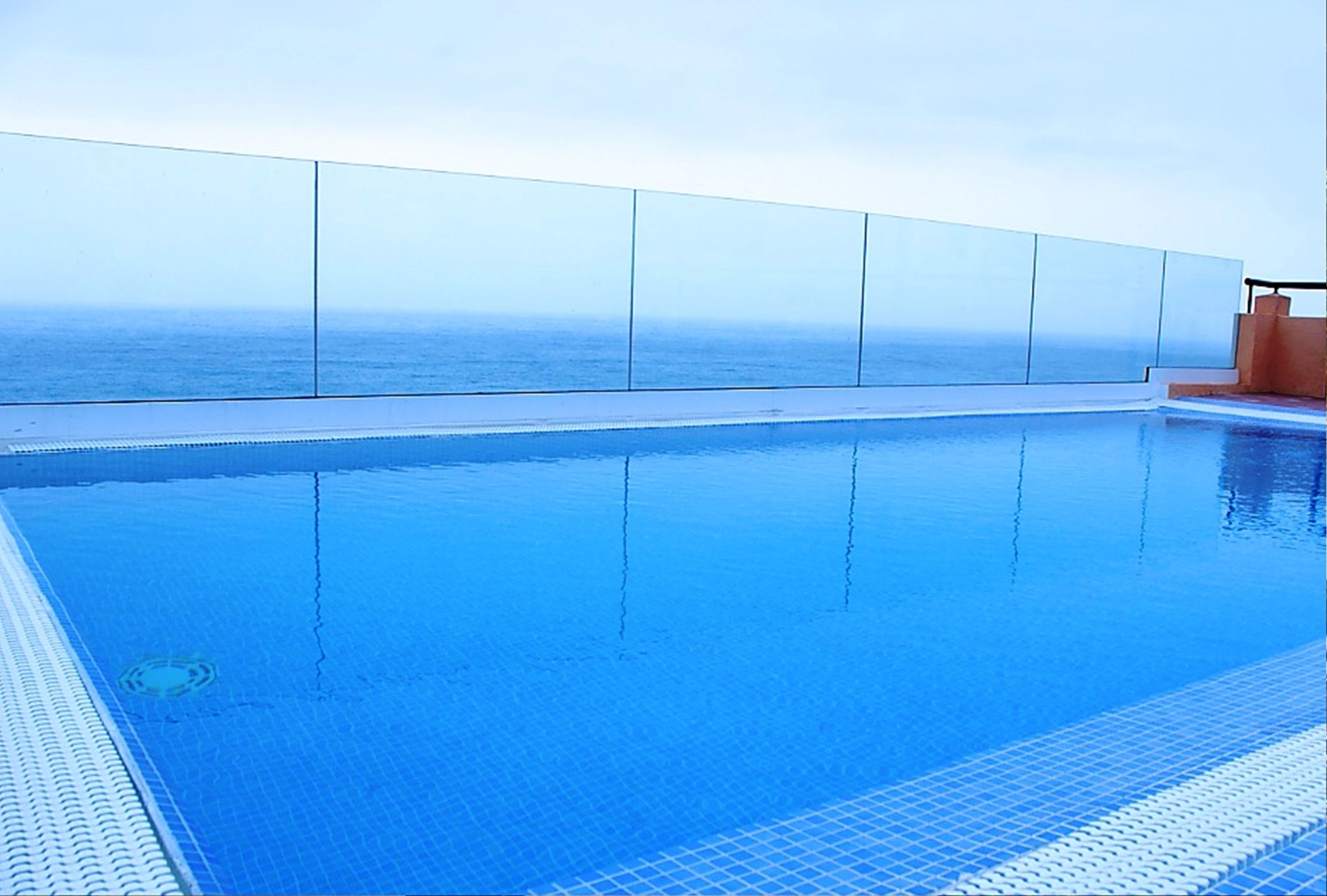 A clear blue pool with a glass barrier and a view of the ocean.