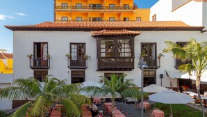 Un edificio con fachada blanca y tejado de tejas rojas, con balcones adornados con plantas en macetas y áreas de estar al aire libre con sombrillas.
