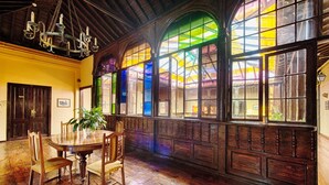 Un comedor con mesa y sillas de madera, ventanas amplias con vidrieras y una araña.