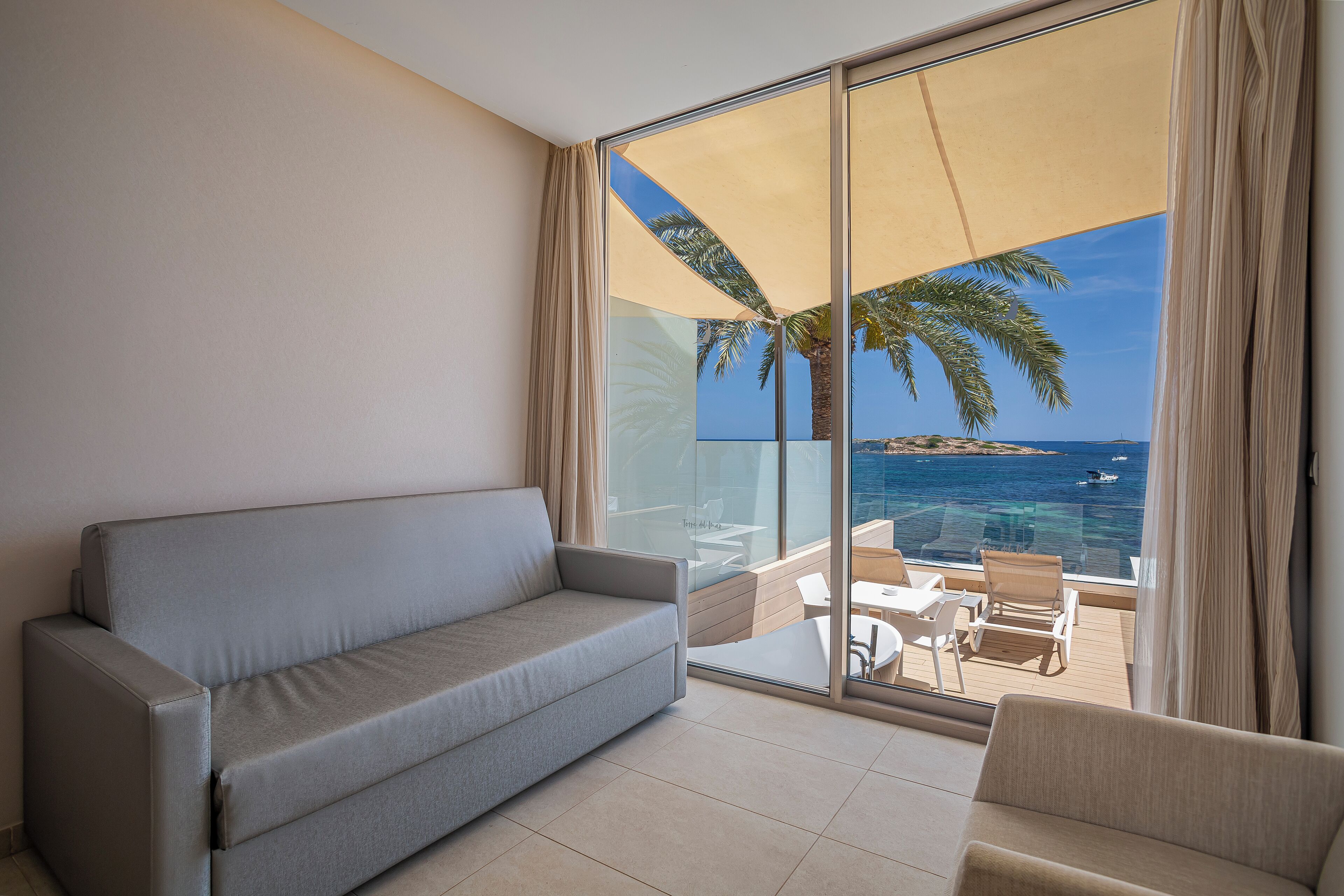 Un balcón con sofá y sillones, un ventanal de vidrio y vistas al mar y palmeras.