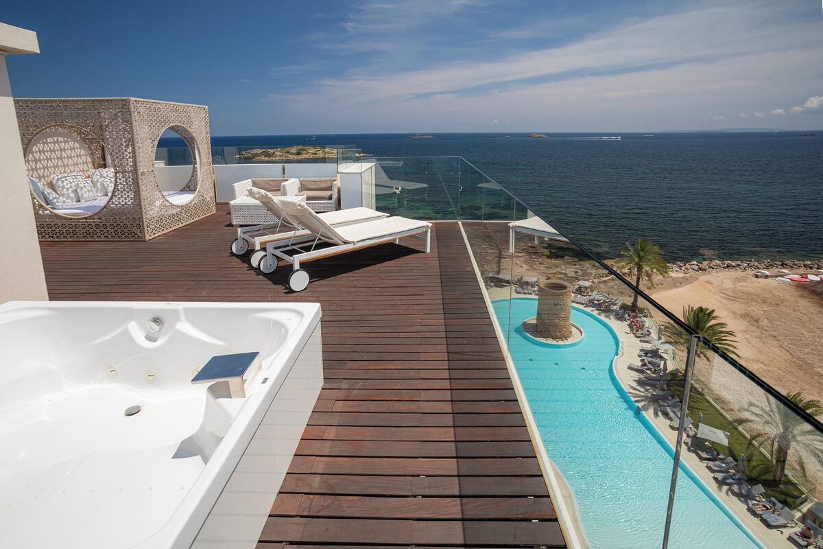 A rooftop pool area with a hot tub, lounge chairs, and a glass balustrade overlooking the sea.