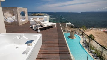 A rooftop pool area with a hot tub, lounge chairs, and a glass balustrade overlooking the sea.