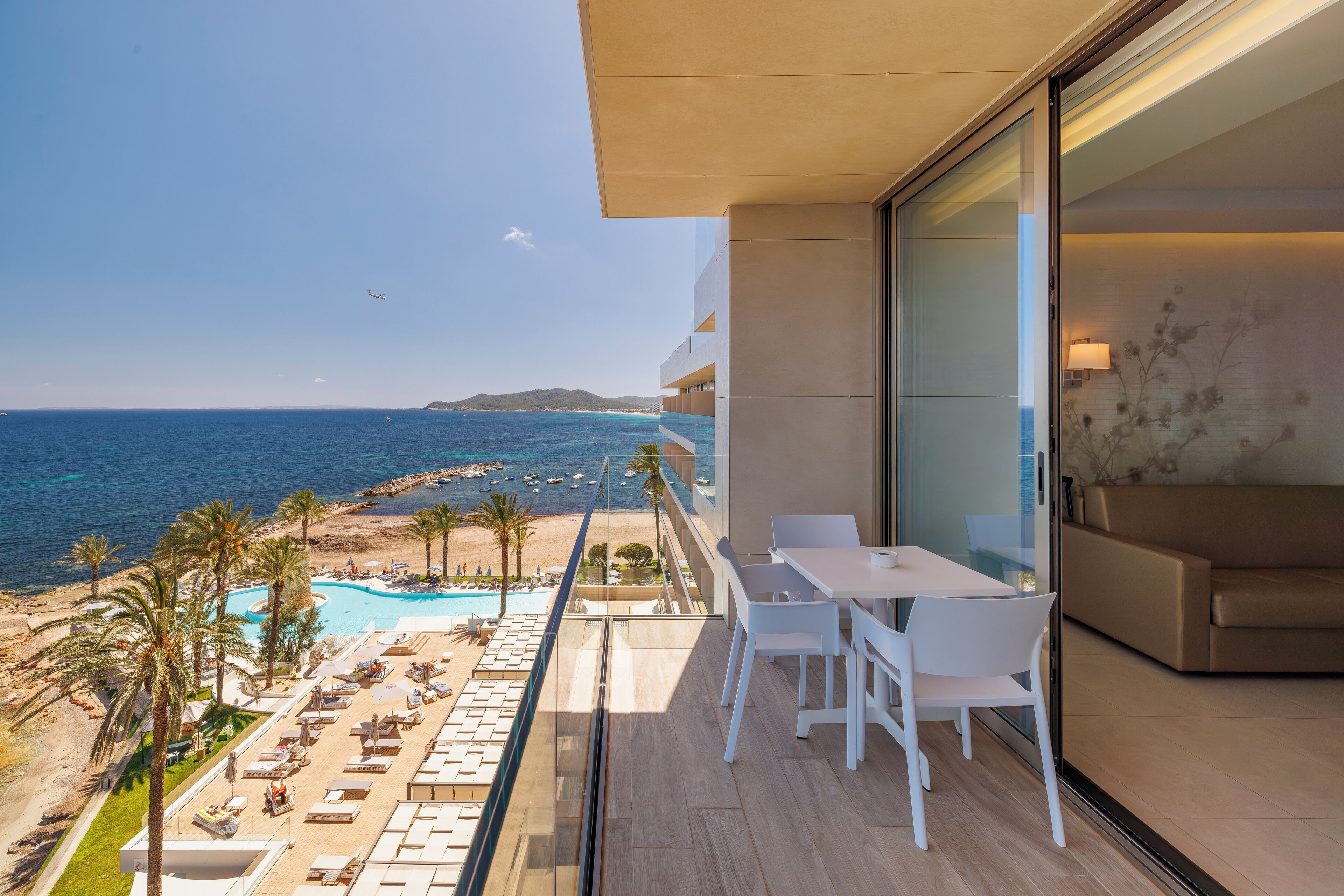 A balcony with a view of the sea, a pool, and a lounge area with chairs and tables.