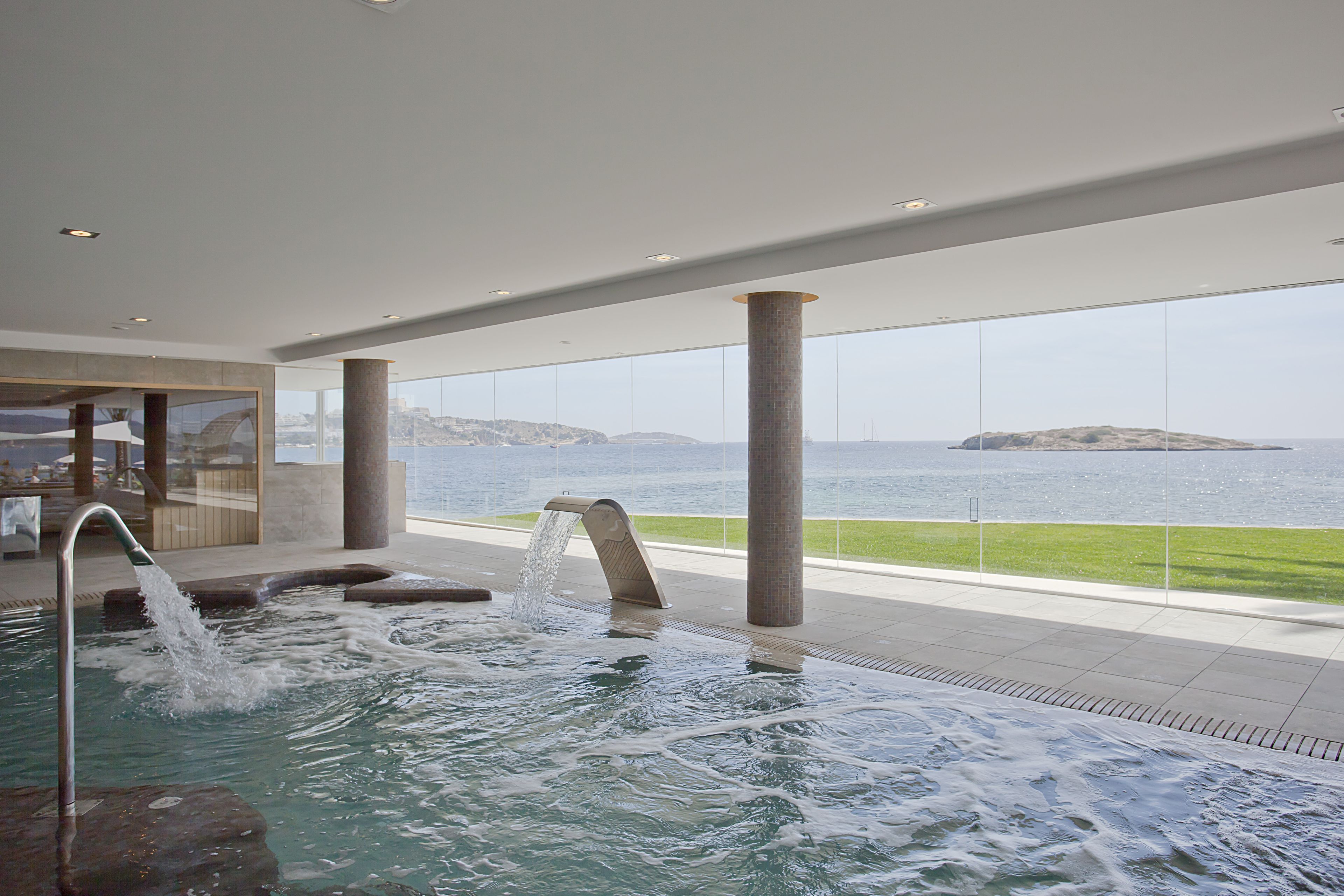 Une piscine intérieure moderne agrémentée d’un élément aquatique décoratif, de grandes fenêtres offrant une vue sur la mer et d’une structure en colonnes de pierre.