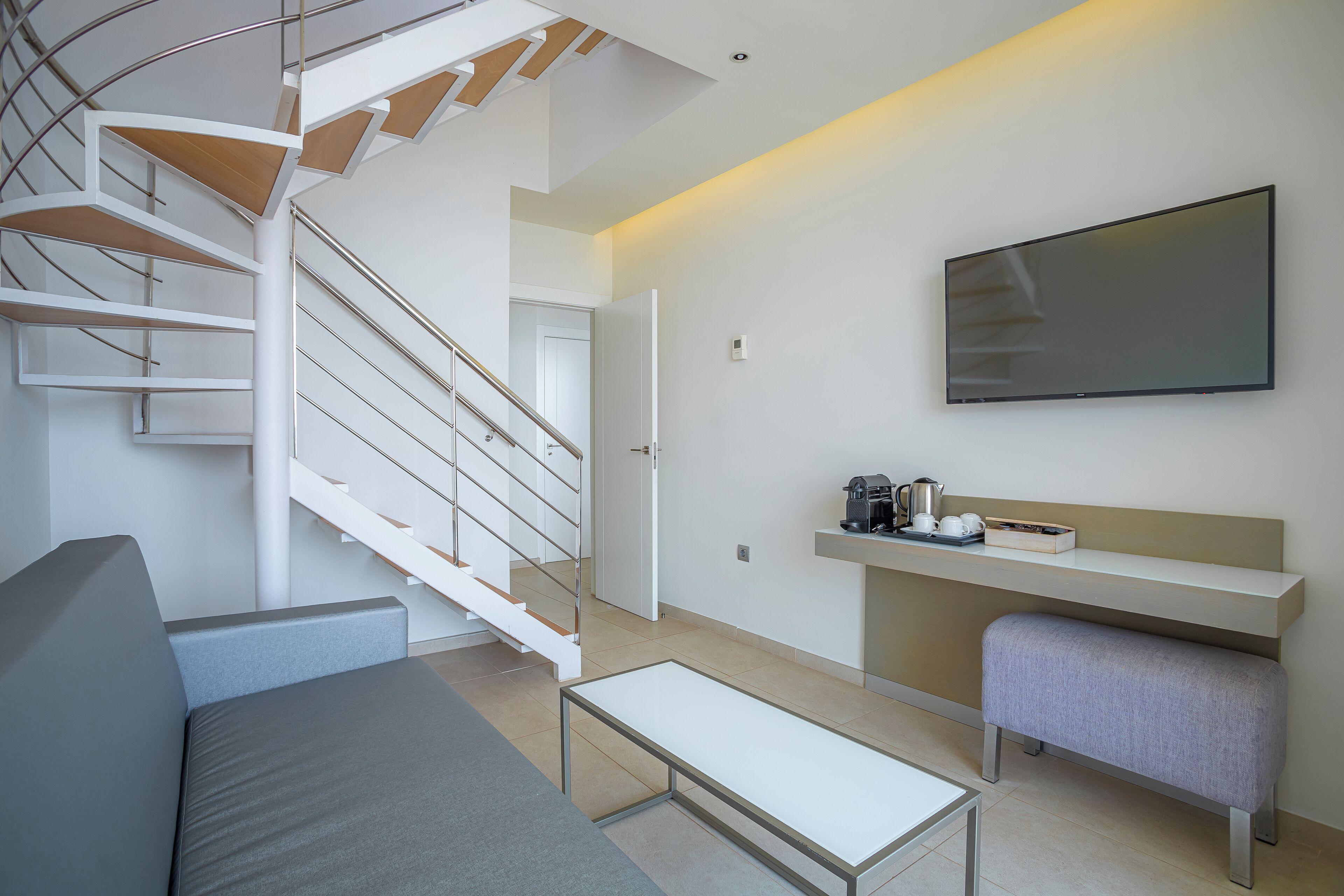 A modern living room with a staircase, a flat-screen TV, a coffee station, and a seating area.