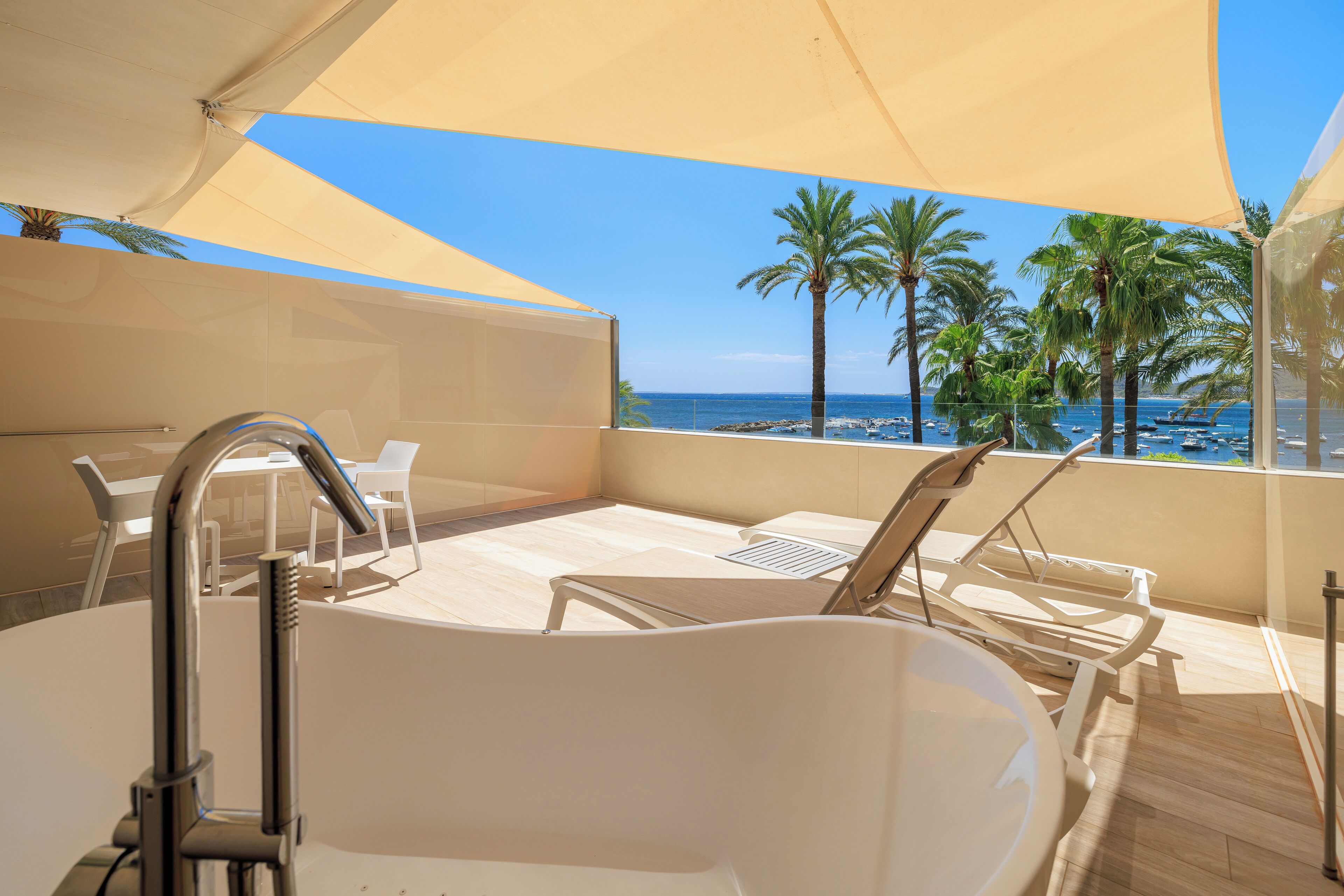 Un balcón soleado con una bañera blanca, sillas de descanso y vistas al mar y palmeras.