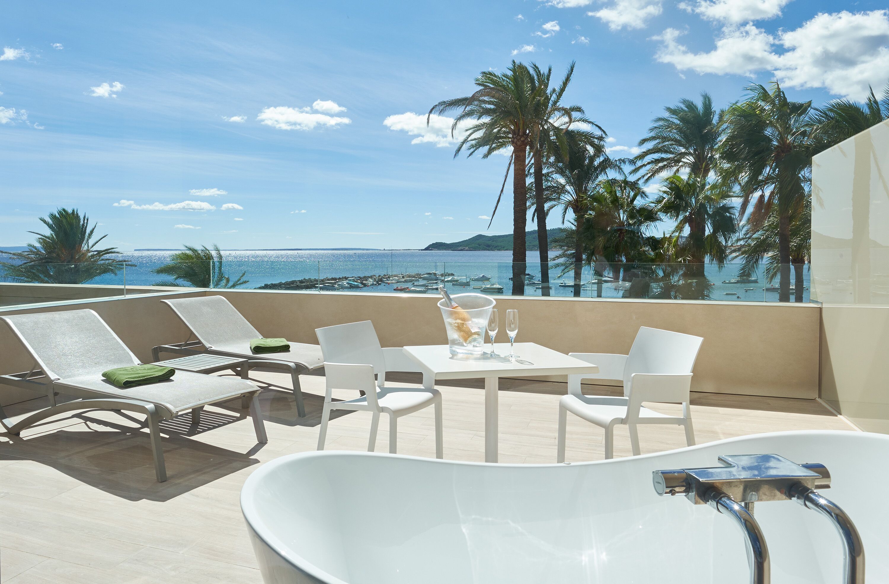 Varanda com vista para o mar, cadeiras de descanso brancas, uma mesa com um copo de água e uma banheira.