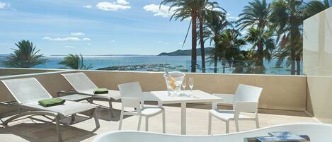 Un balcón con vistas al mar, sillones blancos para relajarse, una mesa con un vaso de agua y una bañera.