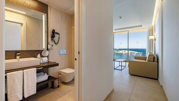 A modern bathroom with a large mirror, sink, and towel rack. A hairdryer is mounted on the wall. The room has a view of the ocean through large windows.