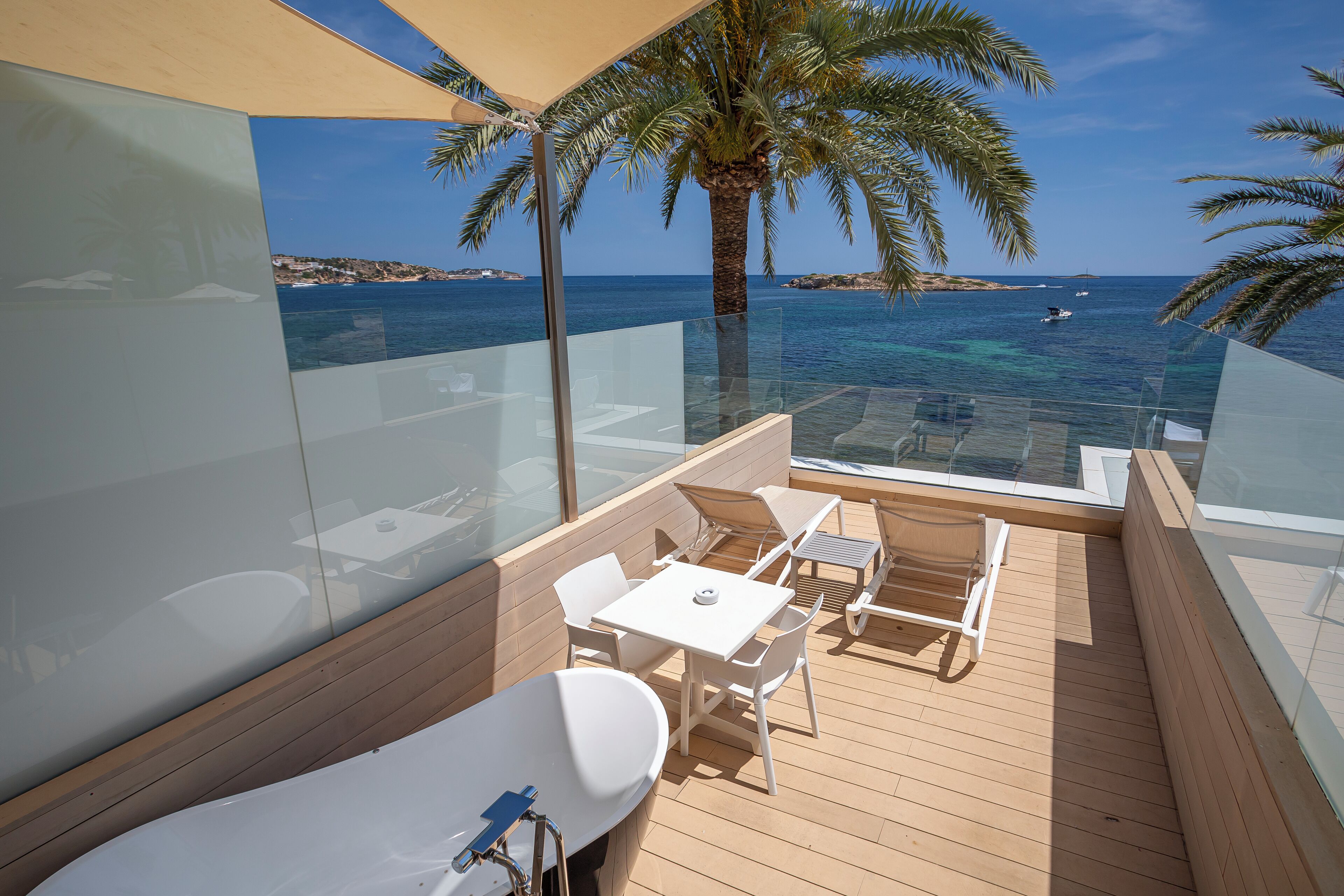 A balcony with a glass railing, white furniture, and a view of the sea and coastline.