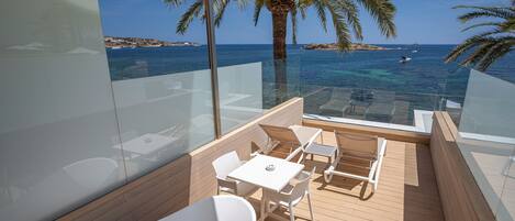 A balcony with a glass railing, white furniture, and a view of the sea and coastline.