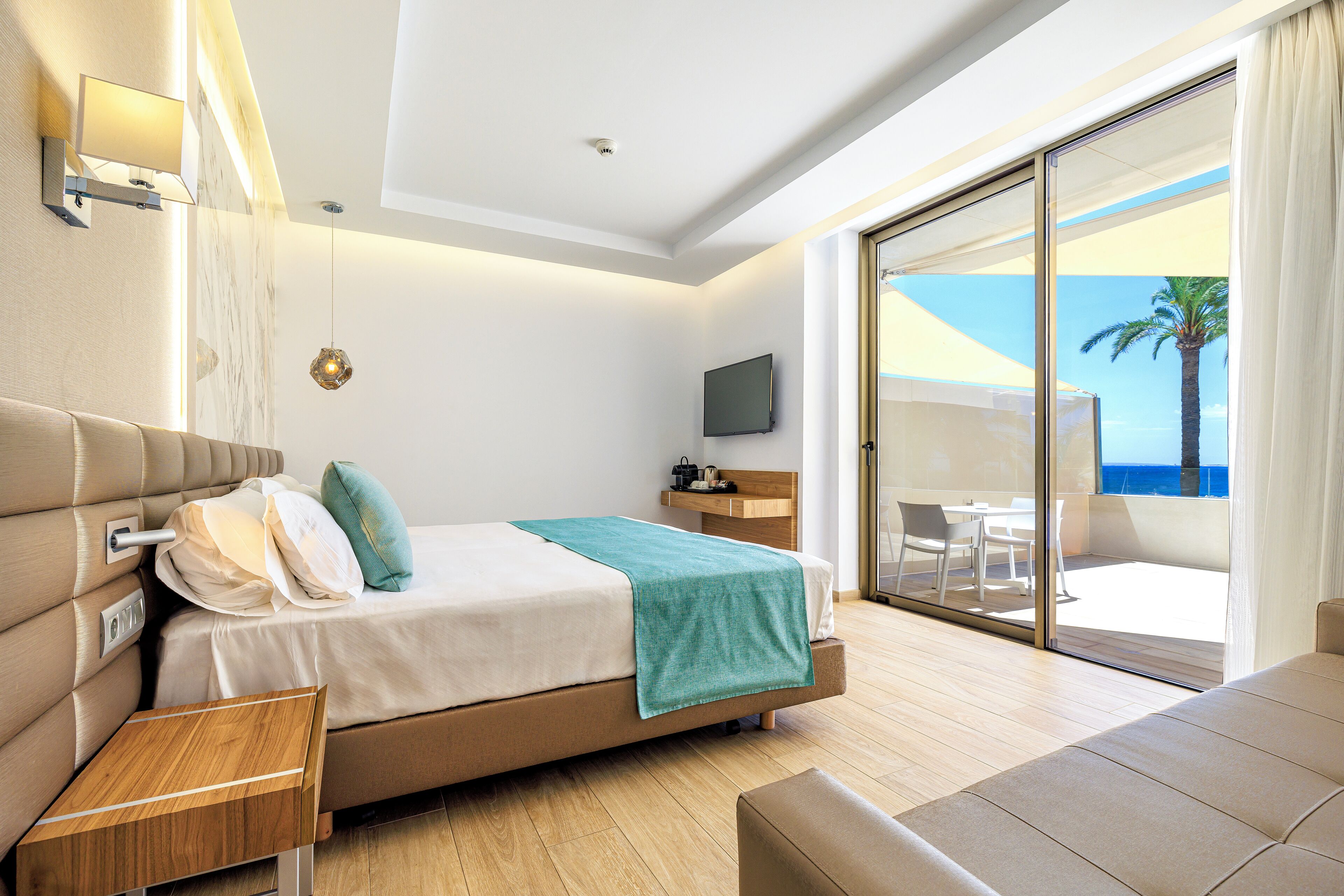 Una habitación de hotel moderna con cama, televisión, mesita de noche de madera y balcón con vista al mar.