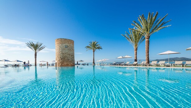 Una piscina con agua cristalina, rodeada de palmeras y sillones de descanso bajo sombrillas.