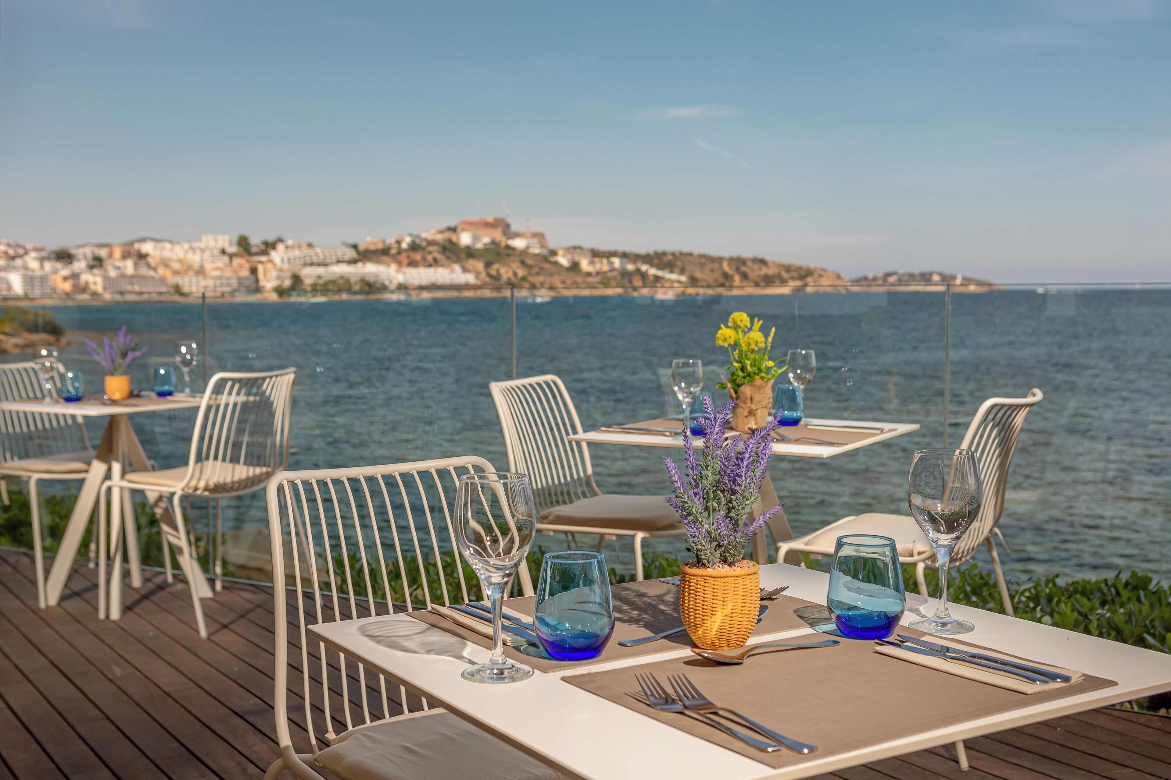 En spiseplads ved havet med borde dækket til to, med blanke glas og en kurvdekoration i midten.