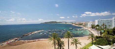 Un hotel frente al mar con vista a la playa, un puerto deportivo con barcos y palmeras.