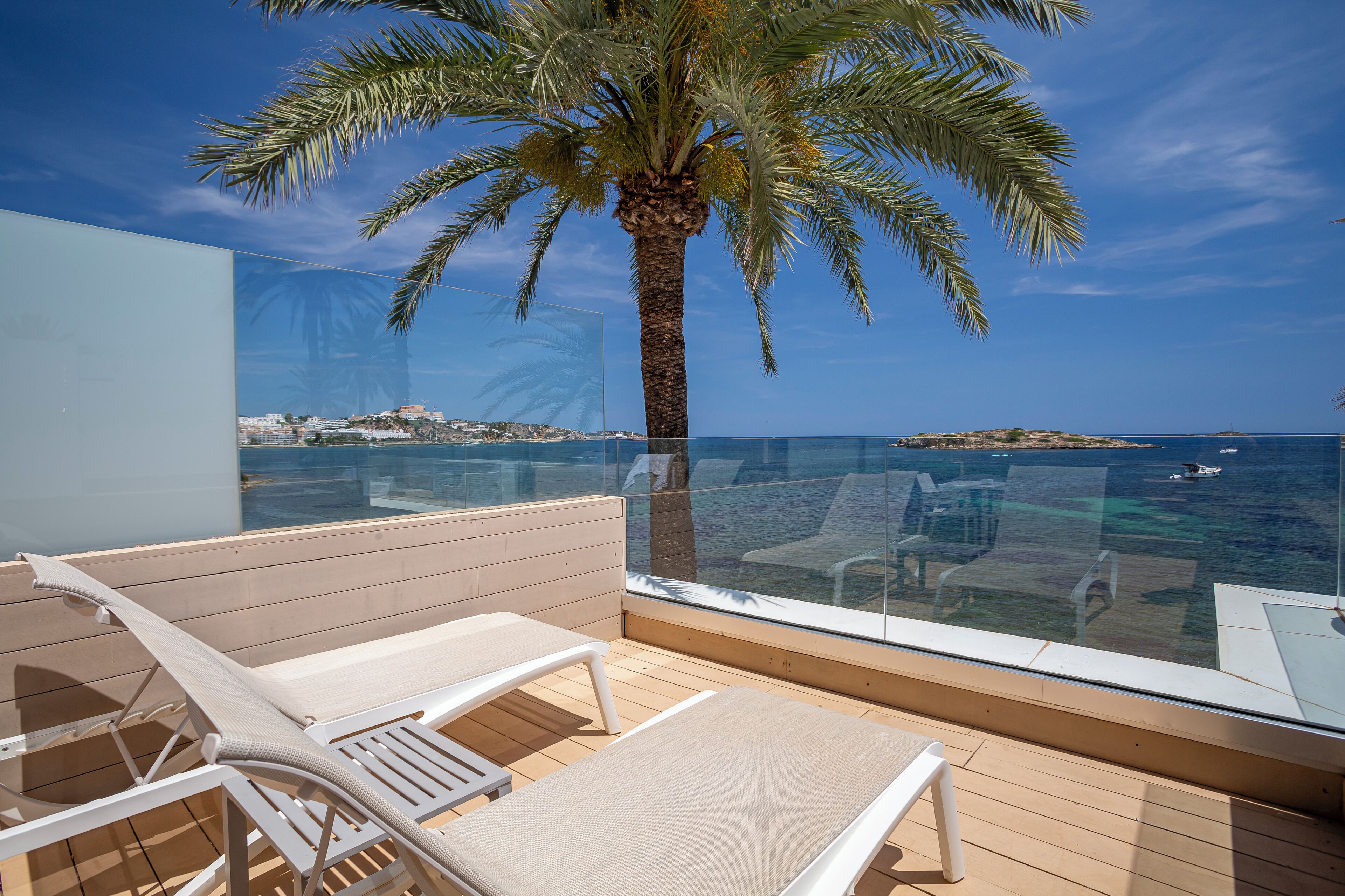 Un balcón con piso de madera, sillones blancos y baranda de cristal con vista al mar.