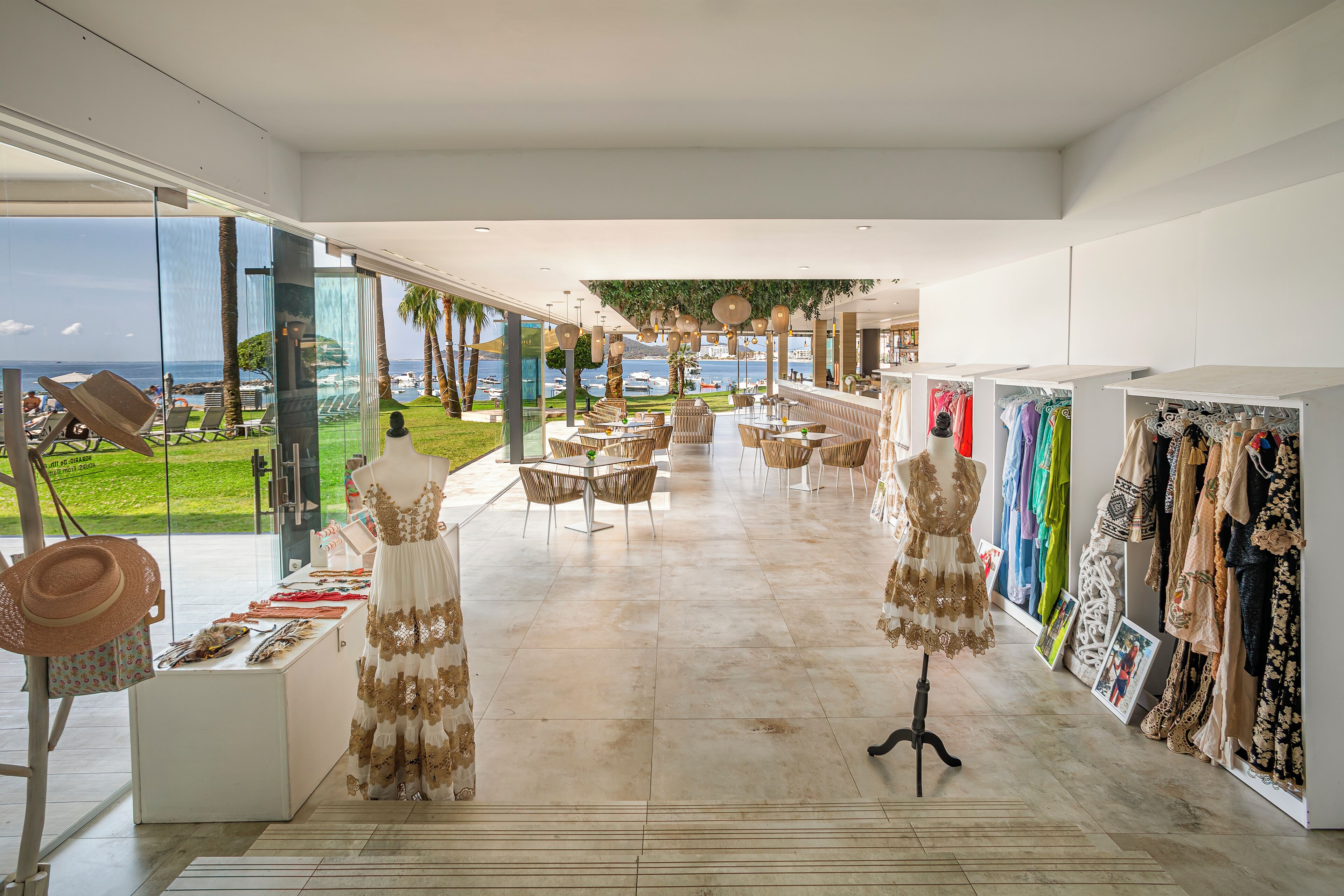 A spacious boutique hotel lobby with a view of the sea, a seating area with tables, and a clothing display.