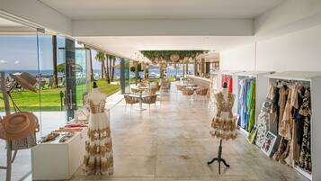 A spacious boutique hotel lobby with a view of the sea, a seating area with tables, and a clothing display.
