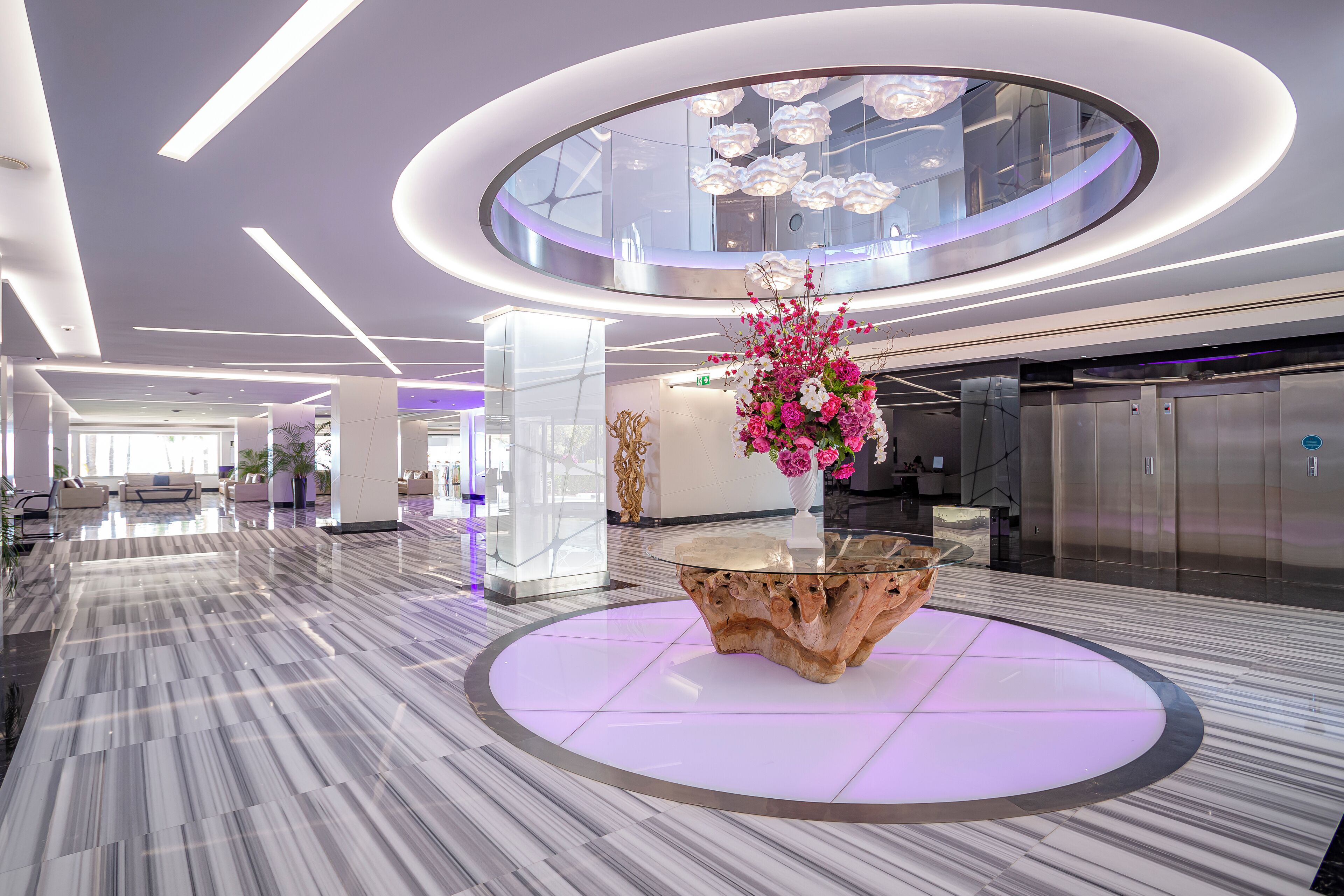 Un hall d’entrée moderne avec un sol en marbre, une table basse en bois placée au centre, agrémentée d’un arrangement floral, et un plafond circulaire orné de luminaires suspendus.