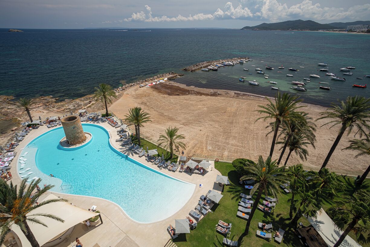 A resort with a large swimming pool, beach, and boats in the background.
