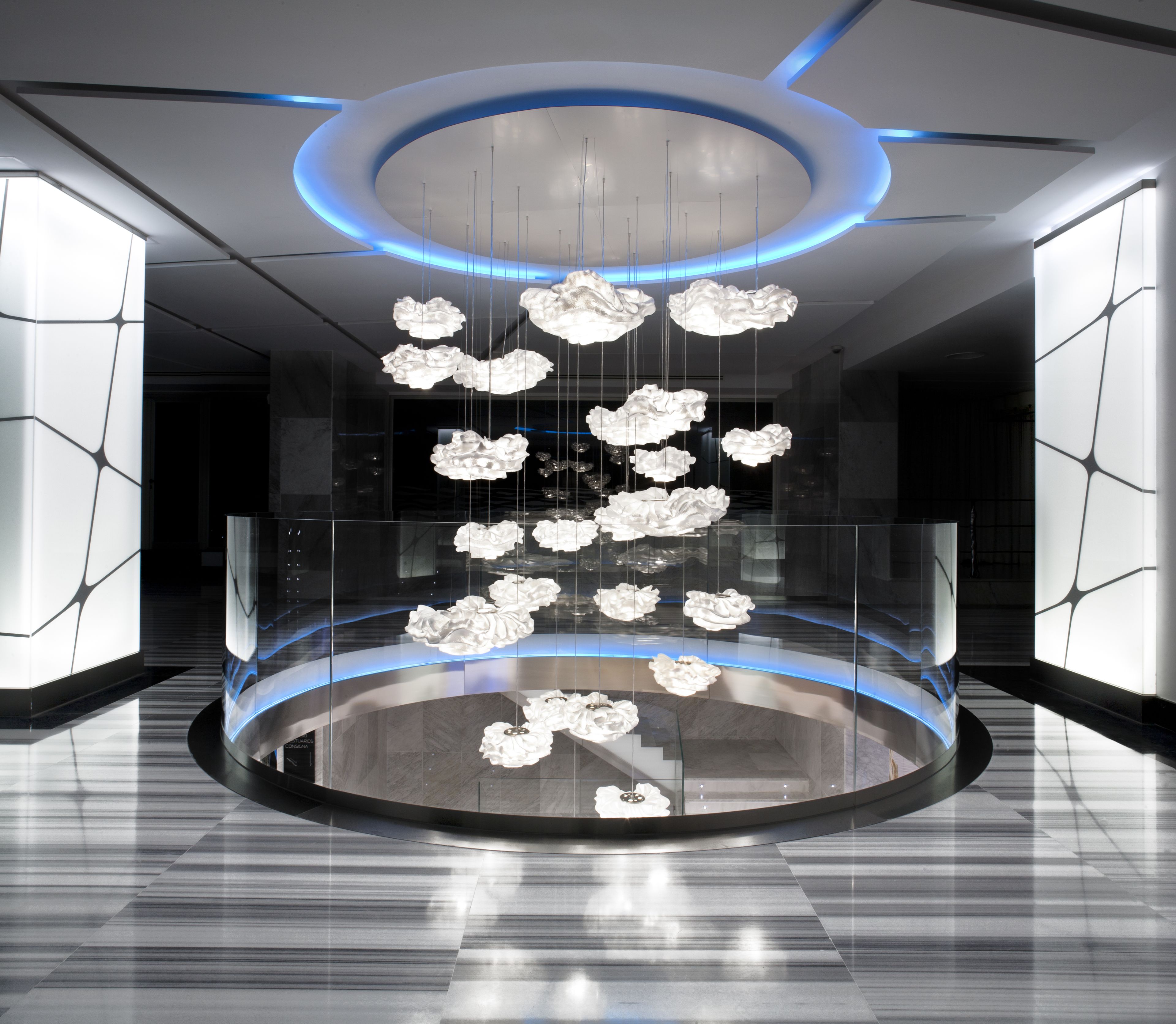 A modern lobby with a circular glass enclosure, hanging white floral light fixtures, and a blue-lit ceiling.