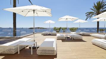 A deck with white lounge chairs and umbrellas, overlooking a body of water with boats and a distant island.