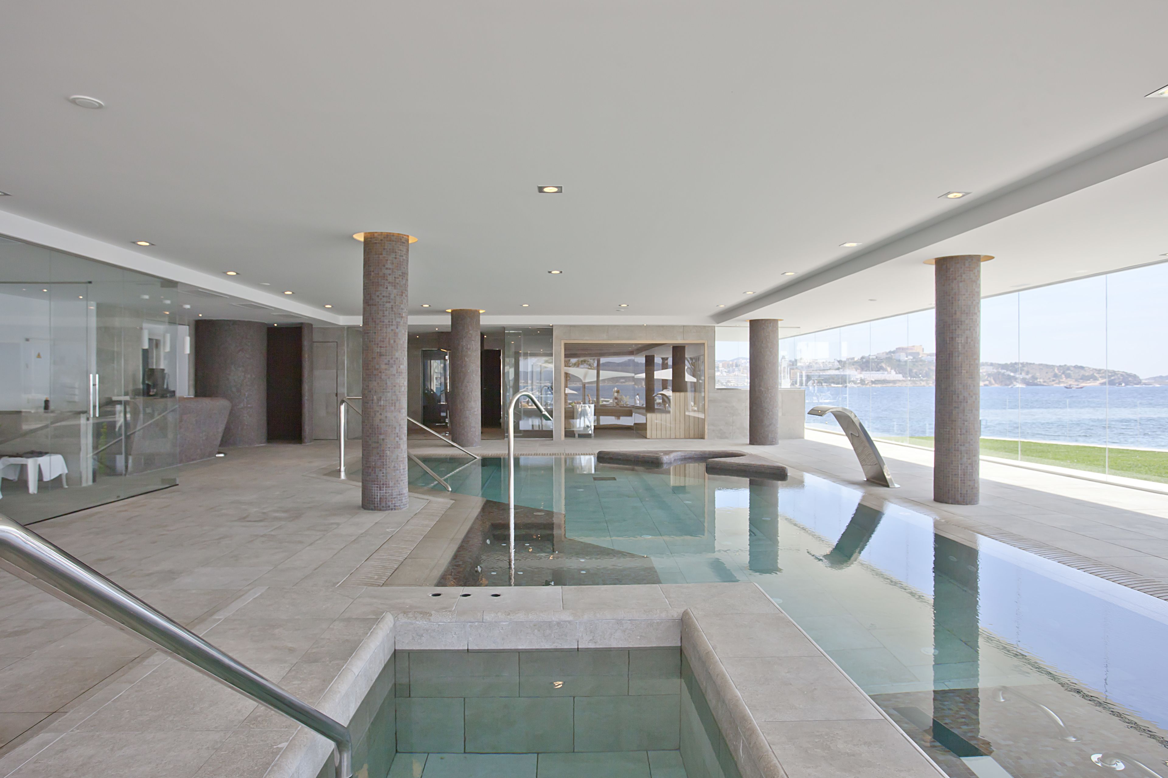 Una piscina interior moderna con paredes de vidrio transparente, pilares grandes y vistas al mar.