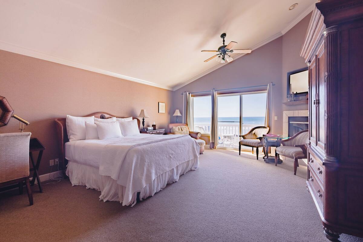 A spacious bedroom with a large bed, a desk, a chair, and a view of the sea through the windows.