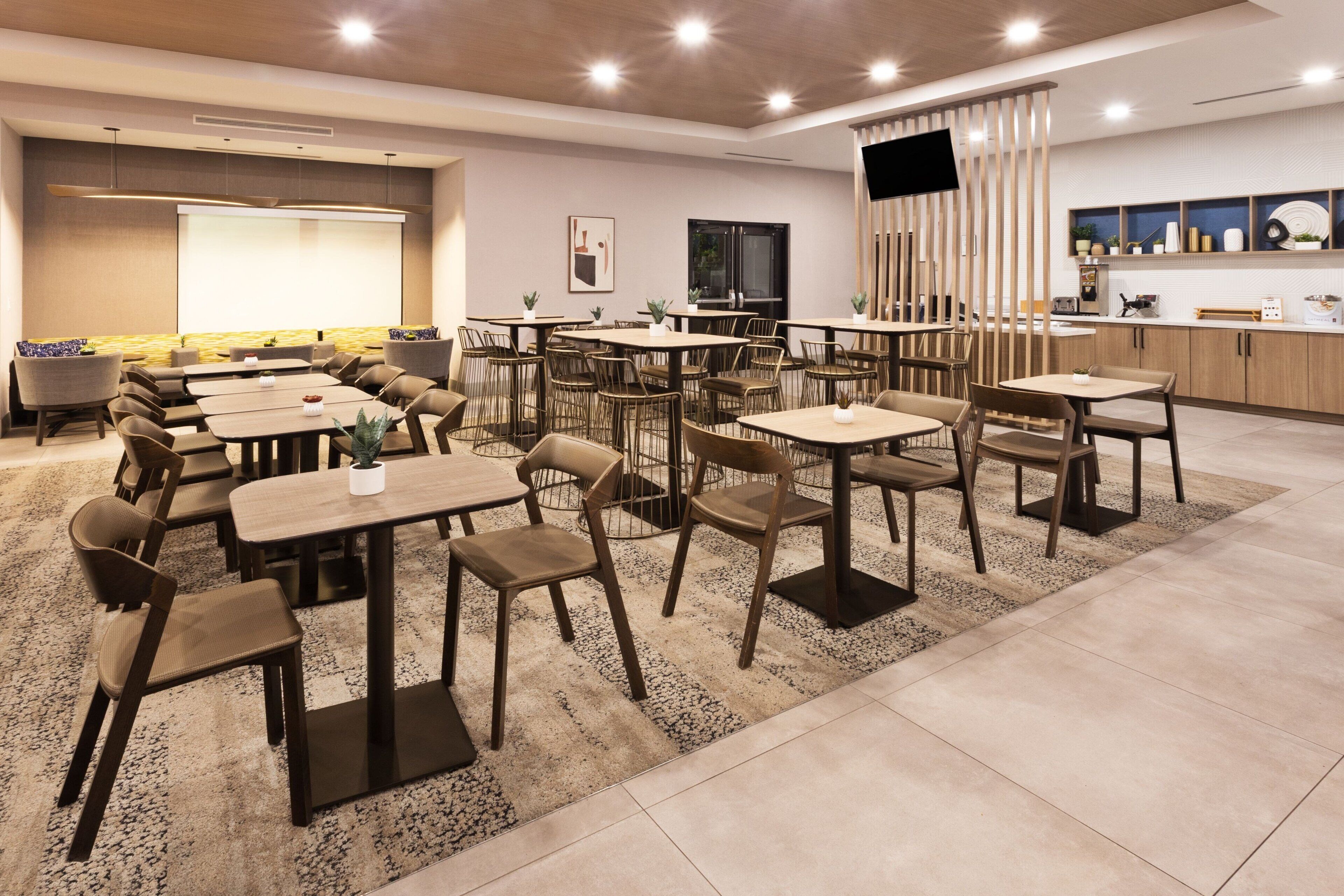 A modern hotel dining area with multiple tables and chairs, a large screen, and a kitchen area.
