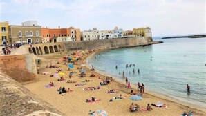 Ein Strand mit badenden Personen, einer Steinmauer und Gebäuden im Hintergrund.