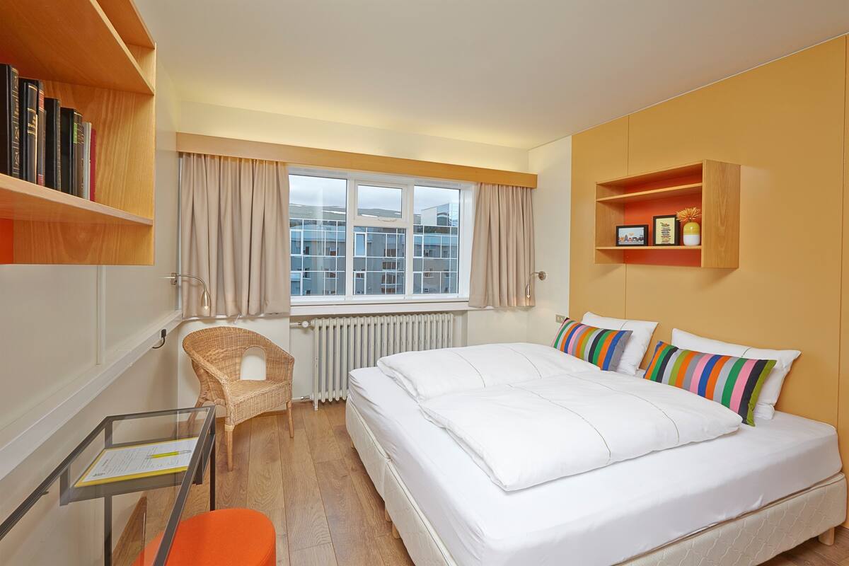 A hotel room with a large bed, a chair, a small table, a window with curtains, and a bookshelf.