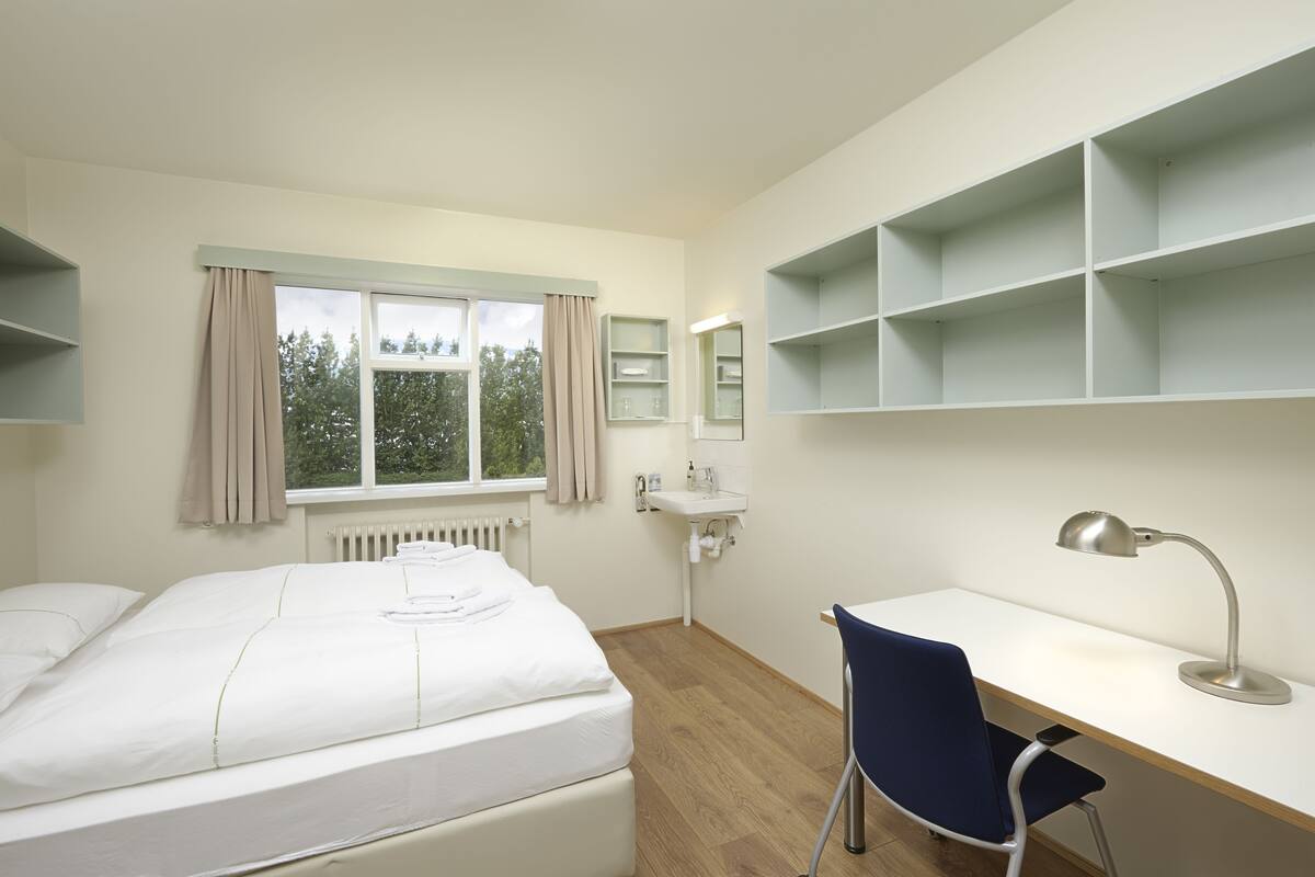 A hotel room with a bed, a desk, a chair, a window with curtains, a radiator, a sink, and shelves.