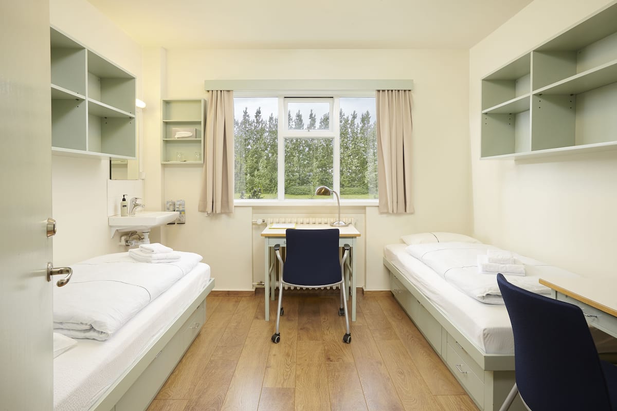 A room with two beds, a desk, a chair, and a window with a view of trees.