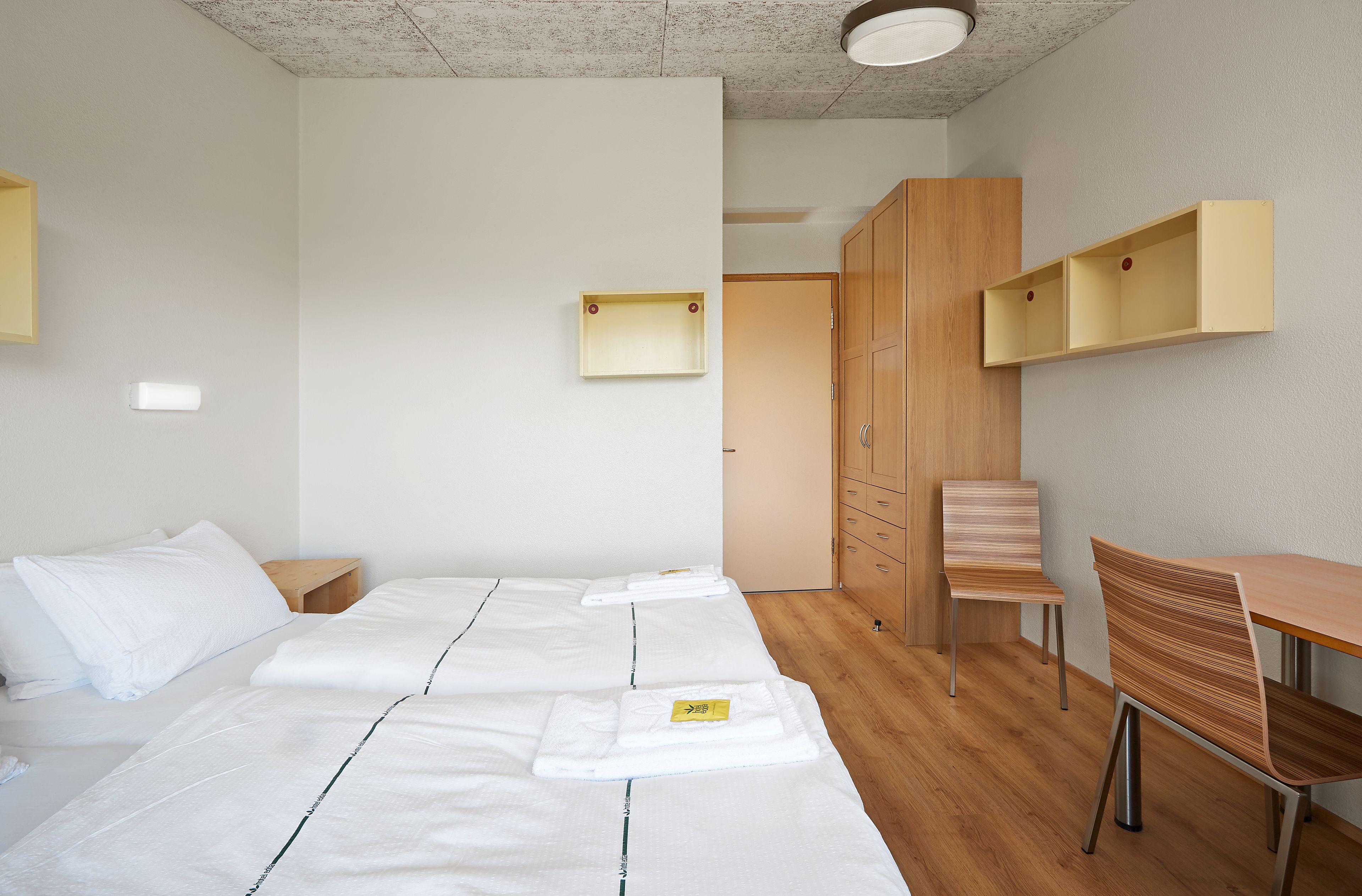 A modern hotel room with a large bed, a wooden desk, and a chair. There is a wardrobe and a shelf unit on the wall.