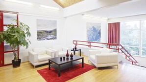 A spacious living area with a wooden floor, white sofas, a coffee table, and a red rug. There are two large windows with red curtains and a staircase with red railings. - Hotel Edda Egilsstadir (Egilsstaðir)