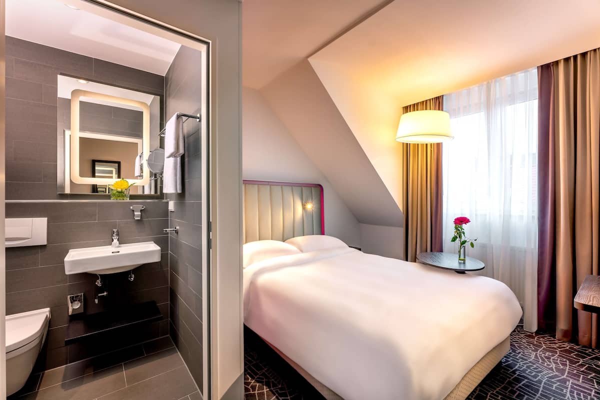 Habitación de hotel moderna con una cama grande, una mesita de noche con lámpara, un baño con lavamanos y espejo, y vistas a la ciudad a través de la ventana.