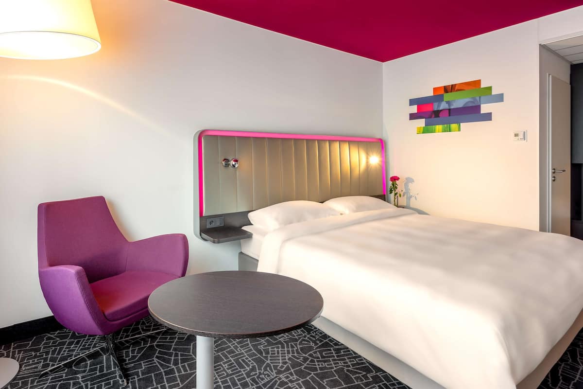 Habitación de hotel con cama, una silla, una mesita pequeña, una lámpara de pared y una obra de arte abstracta y colorida.