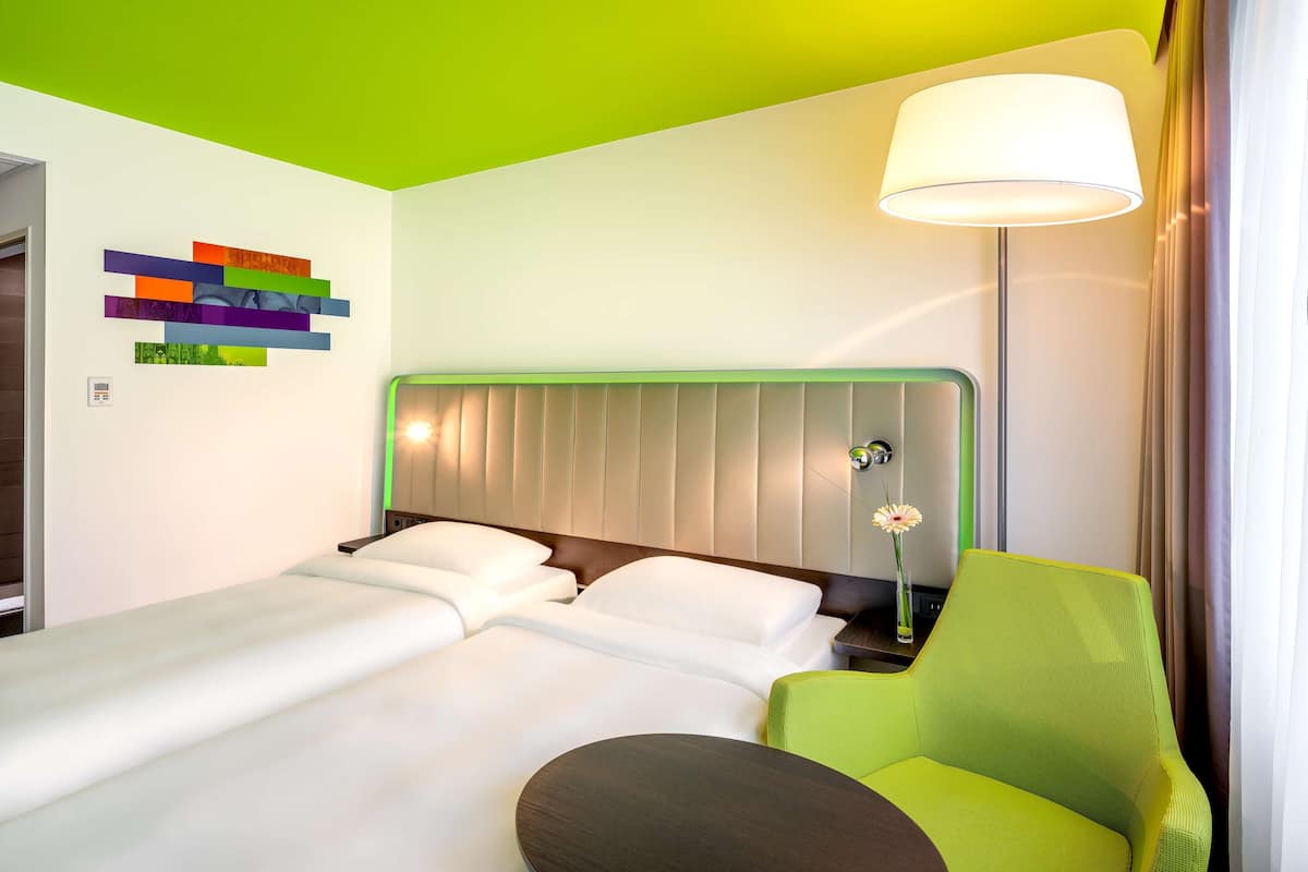 Una habitación de hotel con una cama, una silla verde, una mesita de noche y una lámpara de pared.