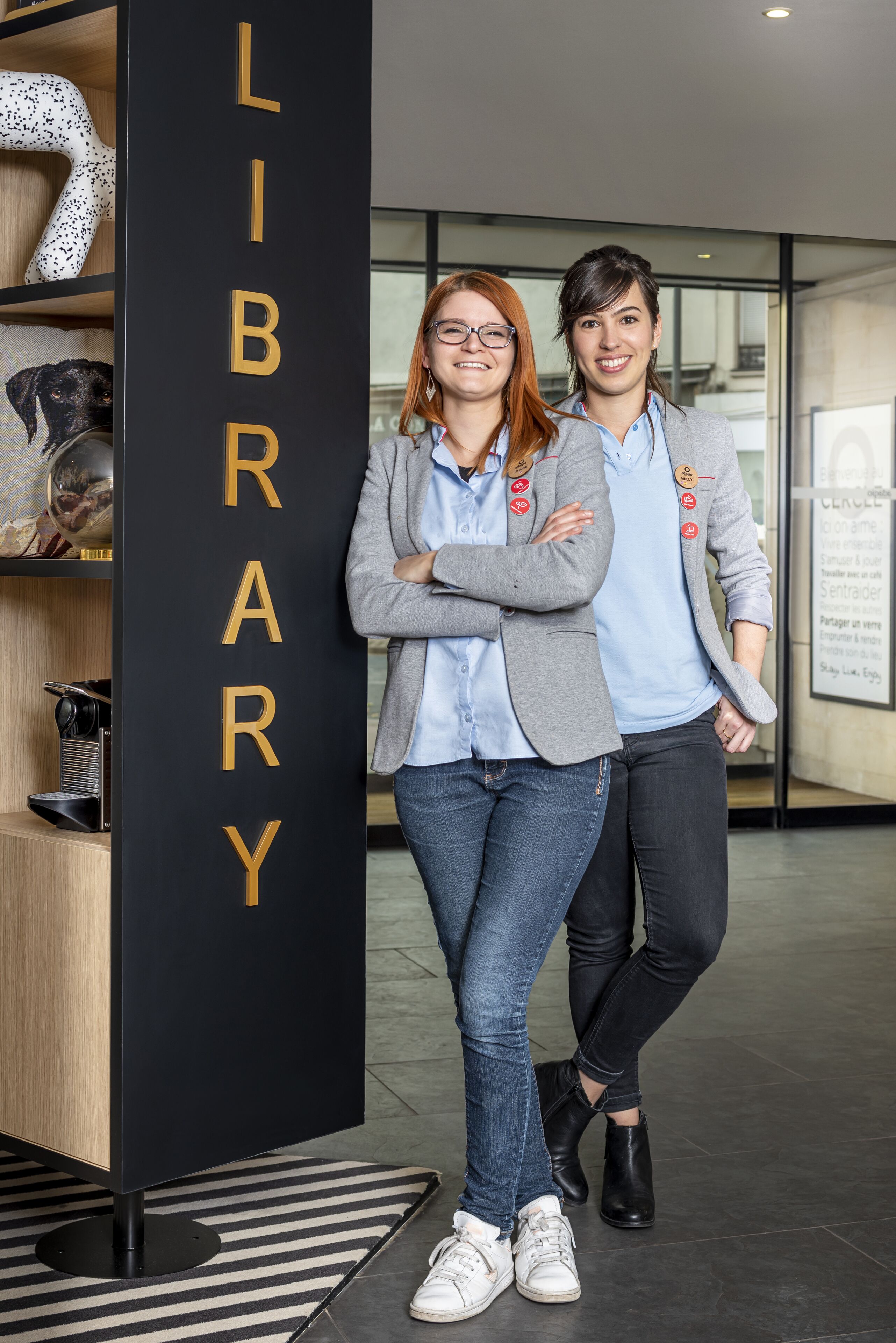 Two people standing in a modern library with a black bookshelf labeled "LIBRARY" and a glass door in the background.