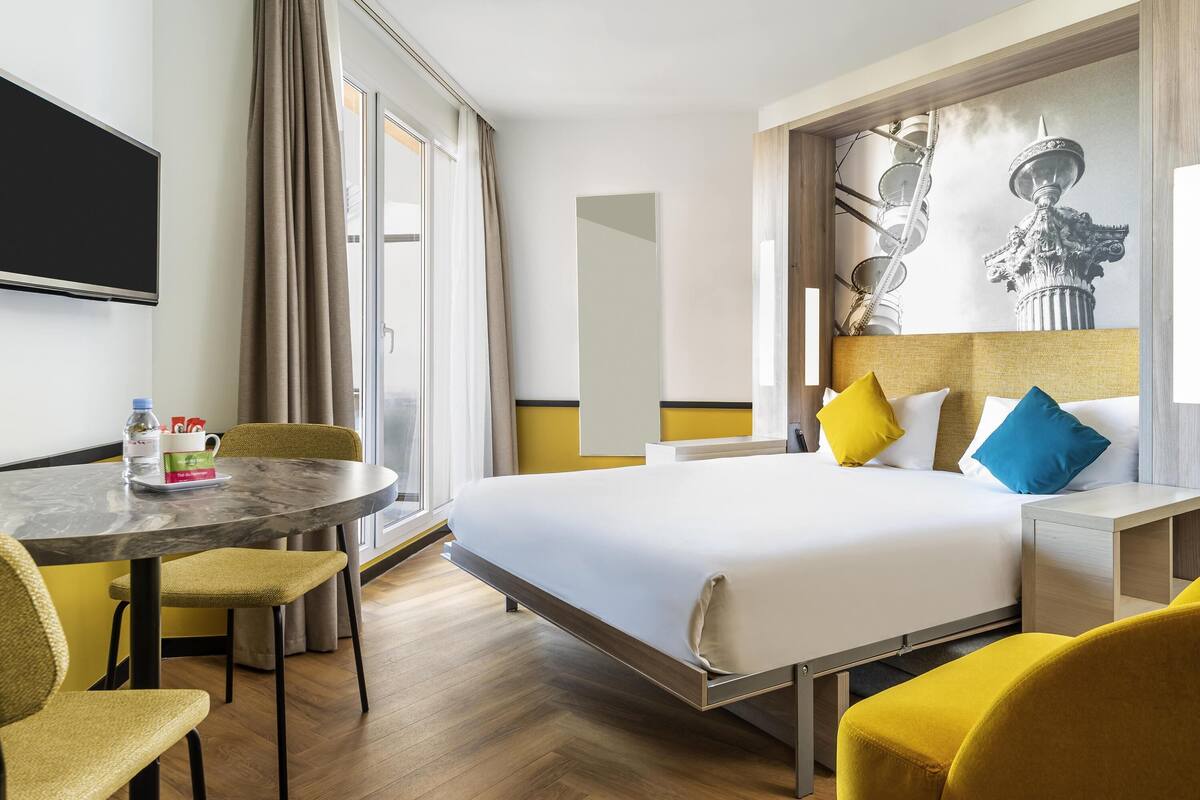 Ein Hotelzimmer mit Bett, Schreibtisch mit Stuhl, Fernseher und einem großen Fenster mit Vorhängen.