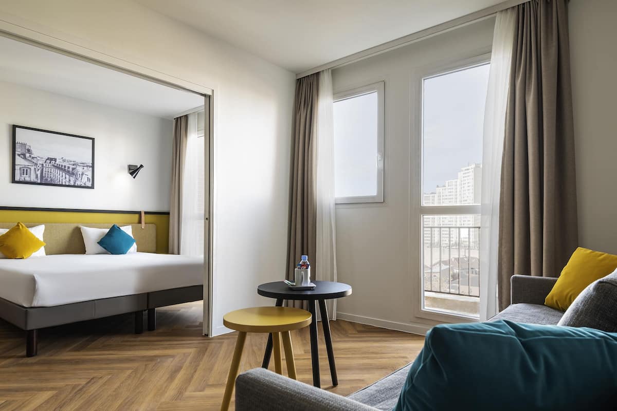 Ein Hotelzimmer mit einem Bett, einem kleinen runden Tisch, einer Sitzecke und Blick auf Gebäude durch die Fenster.