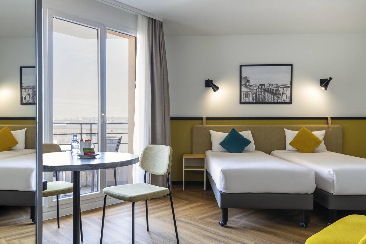 Ein Hotelzimmer mit zwei Betten, einem Esstisch und einem Stuhl. Zwei Wandlampen und ein gerahmtes Bild zieren die Wand.