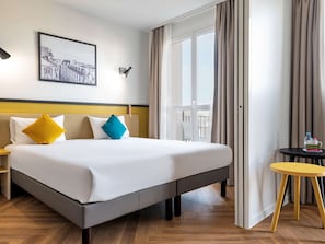 Ein Hotelzimmer mit einem großen Bett, einem Nachttisch, einem kleinen runden Tisch und einem Fenster mit Vorhängen.