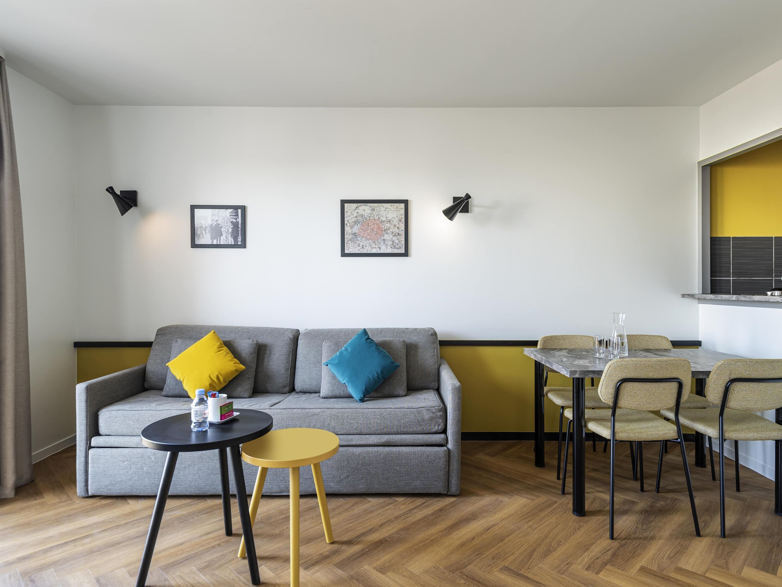 Una sala de estar moderna con un sofá gris, cojines amarillos y azules, una mesita redonda y una zona de comedor con sillas y una mesa.