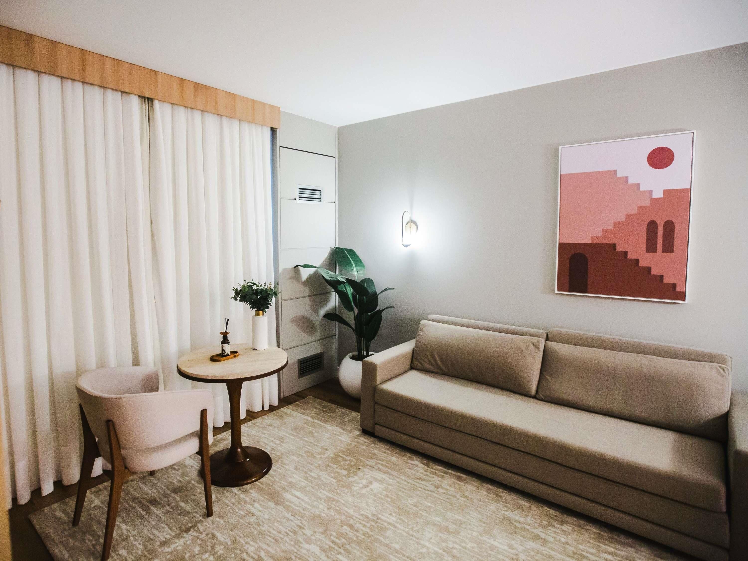 Una sala moderna con un sofá color beige, una mesa redonda y dos sillones. Hay una planta en maceta sobre la mesa y otra sobre el sofá. También se observa una lámpara fijada a la pared y un cuadro enmarcado.
