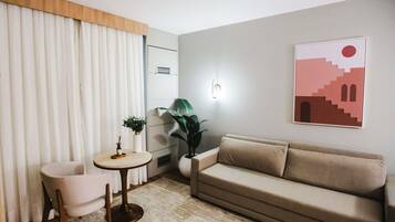 Una sala moderna con un sofá color beige, una mesa redonda y dos sillones. Hay una planta en maceta sobre la mesa y otra sobre el sofá. También se observa una lámpara fijada a la pared y un cuadro enmarcado.