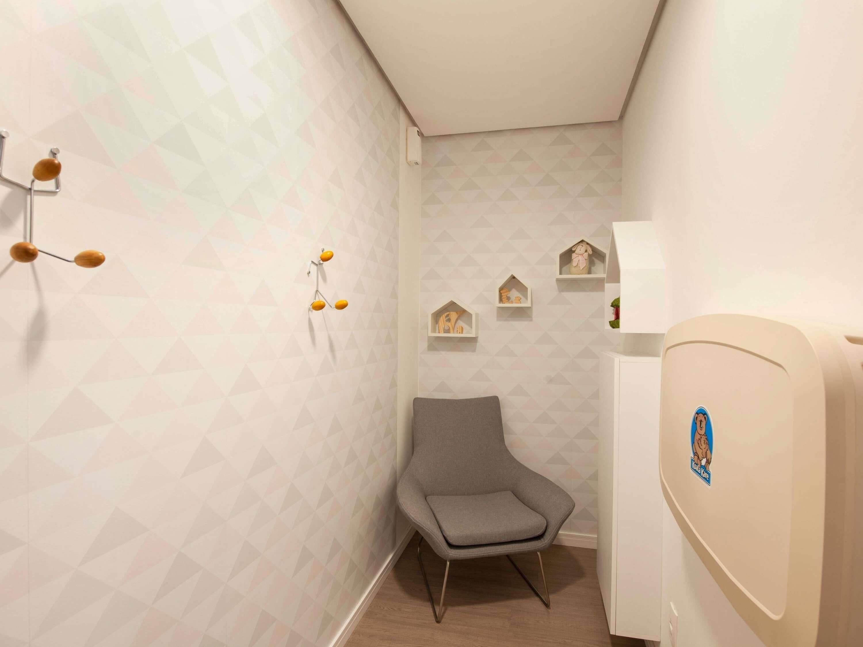A corner room with a grey chair, geometric-patterned wallpaper, a wall-mounted light fixture, and a small shelf with decorative items.