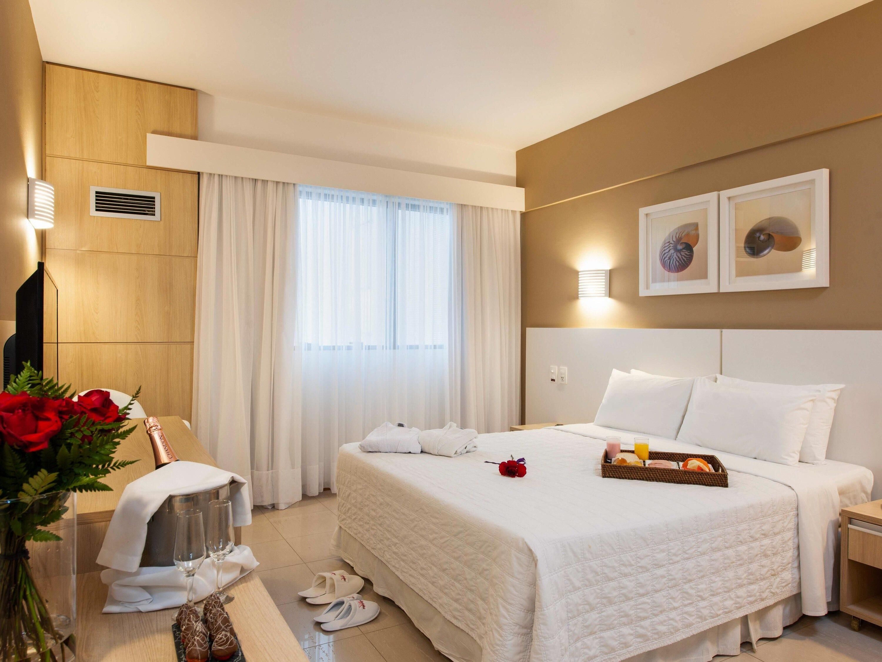 Una habitación de hotel con una cama grande, una mesita de noche con una botella y vasos, un jarrón con flores y una ventana con cortinas.