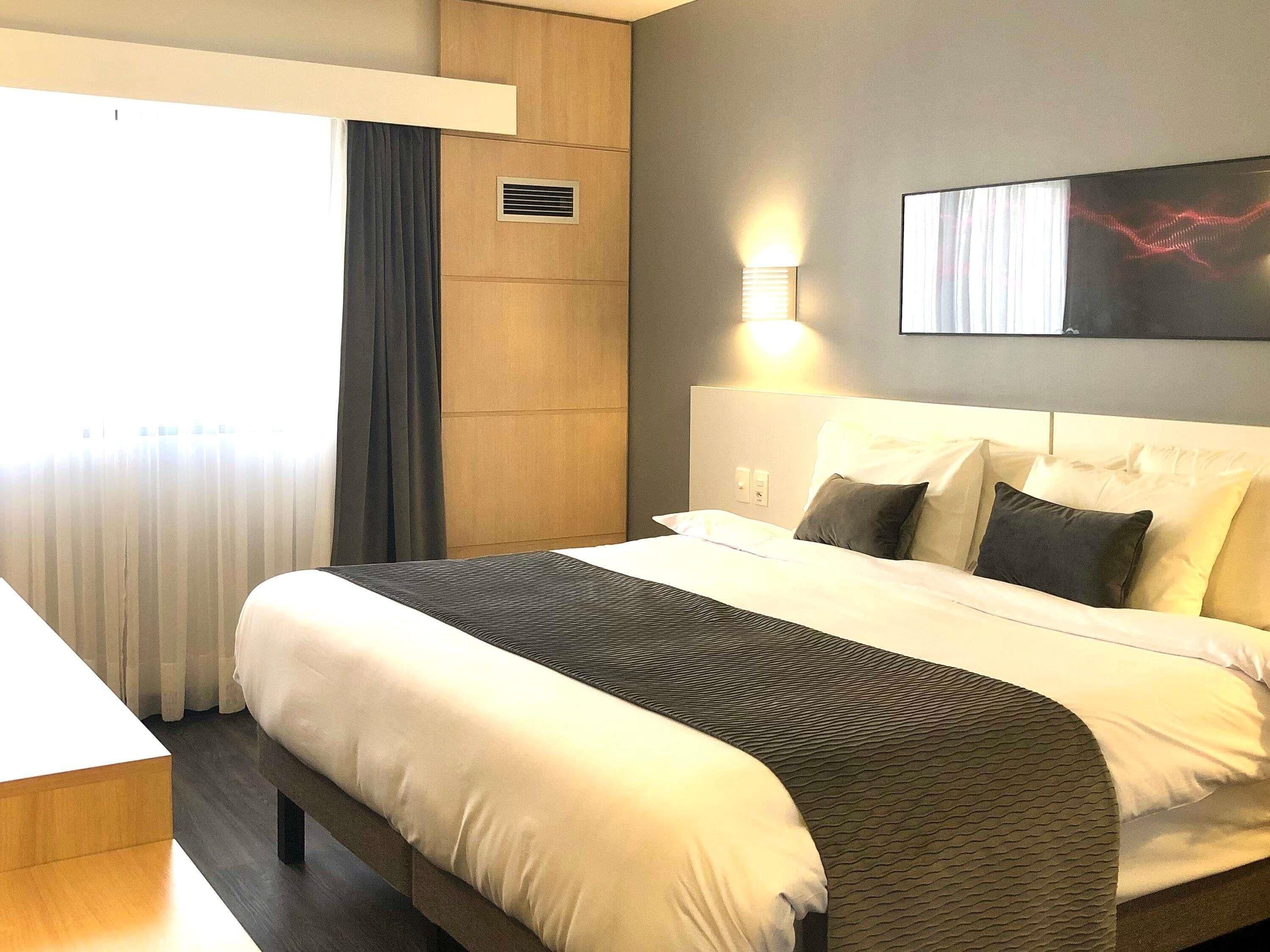 Ein Hotelzimmer mit einem großen Bett, einem Nachttisch, einer Wandleuchte und einem Fenster mit Vorhängen.